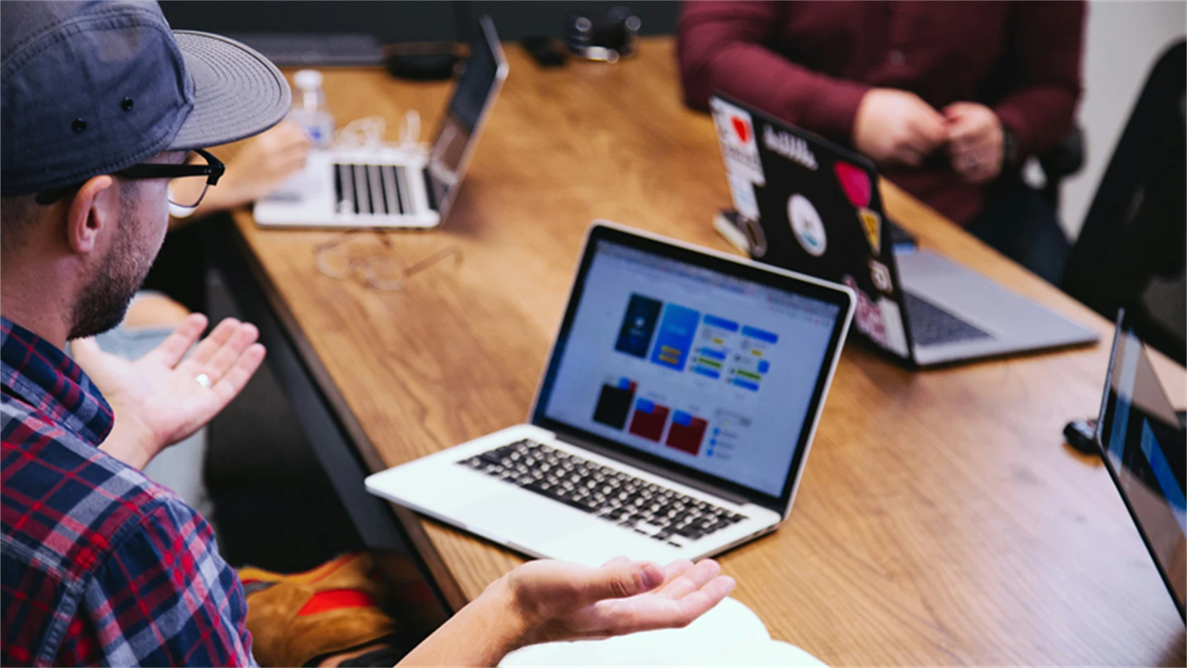 Genrebild på möte mellan personer på en arbetsplats. I förgrunden en man med rutig skjorta och keps på sig med laptop på bordet.